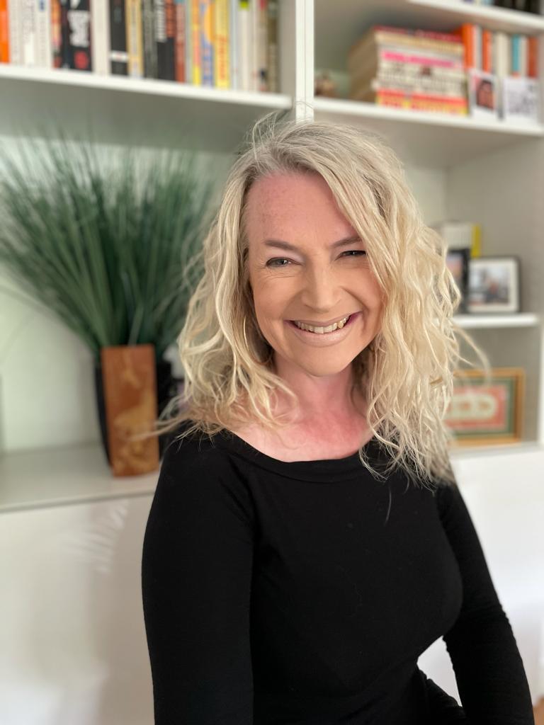 Smiling blonde woman with wavy hair wearing a black top, seated in a bright room with bookshelves behind her.