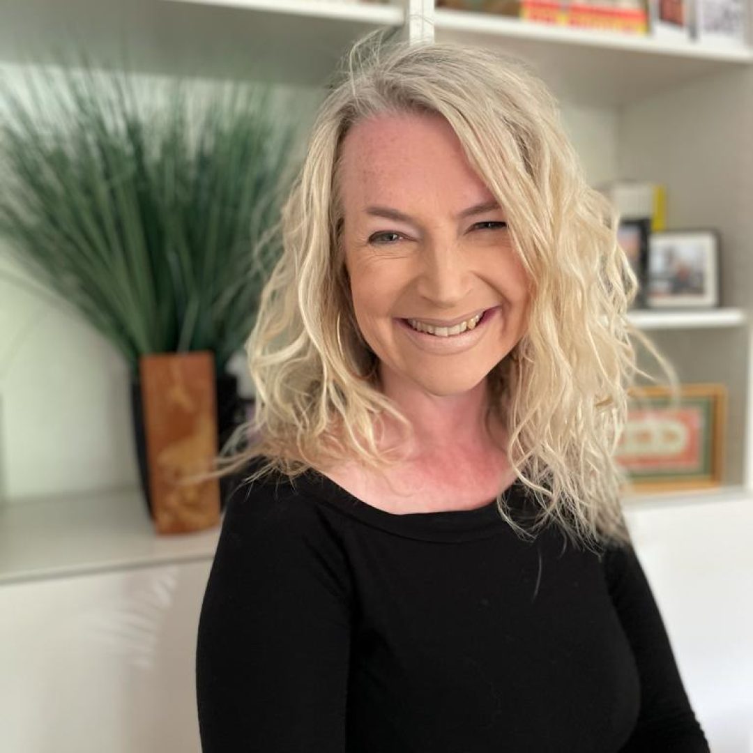 Smiling blonde woman with wavy hair wearing a black top, seated in a bright room with bookshelves behind her.